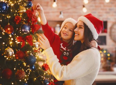 Gülümseyen anne ve kızı evde Noel ağacını süslüyorlar, fiyonk takıyorlar.