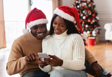 Şenlik Hediyesi Seçiliyor. Noel Baba şapkalı gülümseyen Afrikalı Amerikalı kadın akıllı telefonunu erkek arkadaşına gösteriyor, adam ekranı işaret ediyor, dekore edilmiş oturma odasında aletleri paylaşıyor. İndirim ve Satış