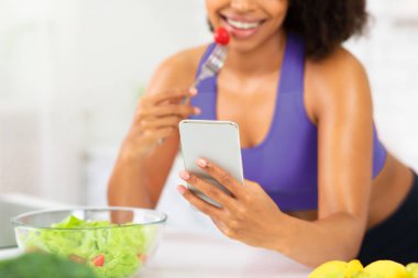 Tanımlanamayan Afrikalı kadın mutfakta salata yiyor, masaya yaslanıyor ve akıllı telefona mesaj atıyor, seçici bir şekilde odaklanıyor.