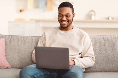 Gülümseyen Afrikalı Amerikalı genç adam evdeki kanepede oturuyor, dizüstü bilgisayar kullanıyor, internetten video izliyor, arkadaşlarıyla sohbet ediyor ya da blog okuyor, kopya çekiyor. Evde Dinlen, Rahatla ve Yaşam Tarzı Konsepti