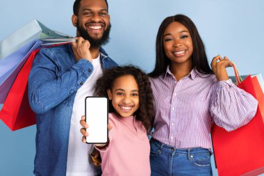 İnternetten alışveriş uygulaması. Elinde alışveriş poşetleri olan mutlu siyah aile portresi, elinde beyaz, izole bir ekranla cep telefonunu gösteren gülümseyen bir kız. Model yapmak için boş alan, mavi stüdyo duvarı.