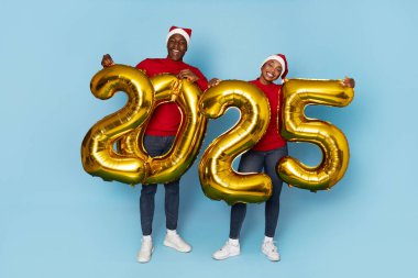 İki siyah arkadaş, 2025 'in gelişini kutluyor. Kırmızı şenlik kıyafetleri ve Noel Baba şapkaları giyiyorlar. Gururla büyük altın sayılar tutuyorlar, neşeli ve neşeli bir atmosfer yaratıyorlar..