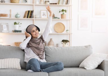 Neşeli bir gün. Evde kablosuz kulaklıkla müzik dinleyen, rahat koltukta dinlenen, boşluğu kopyalayan tesettürlü olumlu Arap Kız