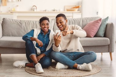 Afro-Amerikan bir çiftin portresi. Halıda oturuyorlar, akıllı telefon kullanıyorlar. Gülümseyen adam aletini tutuyor ve ekranı işaret ediyor, fotoğrafları gösteriyor, sosyal medya içeriğini paylaşıyor.