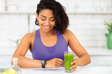Sağlıklı Doğal Yaşam Tarzı. Spor sütyenli gülümseyen Afrikalı kadının portresi. Elinde smoothie, bilek akıllı saatine bakıyor.
