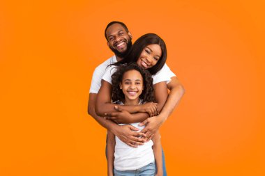 Sağlıklı Aile İlişkisi. Arkasından karısına ve kızına sarılan neşeli Afro-Amerikalı adamın portresi sarı turuncu stüdyo duvarında dikilip kameraya bakıyor.