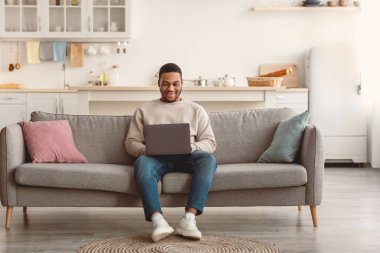 Modern Uzaktan İş Yaşam Tarzı. Evdeki kanepede dizüstü bilgisayar kullanarak gülümseyen genç adam internette çalışıyor, internette geziniyor ya da bilgisayardan video seyrediyor. İnsanlar ve Teknoloji