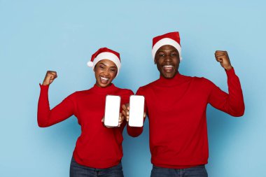 A joyful black couple, wearing matching red sweaters and Santa hats, proudly holds up their smartphones while smiling enthusiastically against a light blue background. clipart