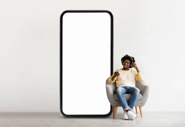 Genç siyah bir adam bir sandalyede rahat bir şekilde oturur ve müzik dinlerken akıllı telefonunun tadını çıkarır. Yanında büyük boş bir ekran duruyor, minimalist bir atmosfer yaratıyor..