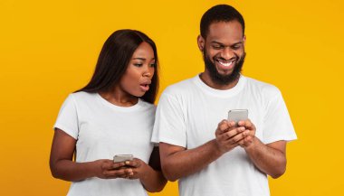 Şok olmuş, gülümseyen erkek arkadaşını gözetleyen Afrikalı Amerikalı bir kadının portresi. Cep telefonu kullanan, mesaj atan ya da sosyal medyadan haber alan. Sarı stüdyo arkasında duran siyah çift.