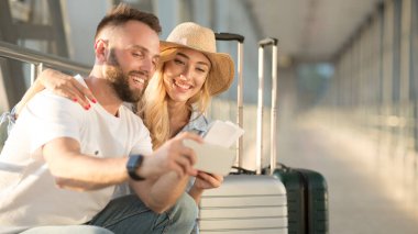 Tatil başlıyor. Mutlu çift biletlerle selfie çekiyor, havaalanı binasında uçak bekliyor.
