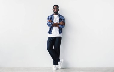 Kollarını kavuşturmuş, kameraya bakıp beyaz duvara gülümseyen gündelik giysili Afro-Amerikalı çekici genç bir adam. Pozitif ifade eden, uzun boylu, havalı siyahi bir adam.