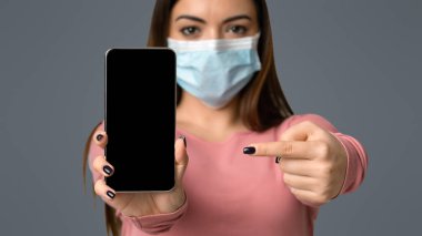 Tıbbi Maskeli Genç Kadın Siyah Ekranlı Akıllı Telefonu Gösteriyor, Işık Stüdyosu Arkaplanında Duruyor, Seçici Odaklanma, Mokup