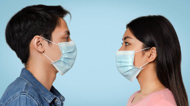 Pandemia Riski. Koruyucu tıp maskeleri takmış genç Asyalı bir çiftin yan görüntüsü. Birbirlerine bakıyorlar, arka planda duruyorlar.