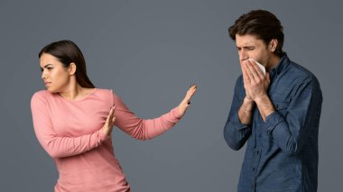 Social Distancing. Young woman disgusted by her sick boyfriend with runny nose, afraid of coronavirus spread, light studio background with free space clipart