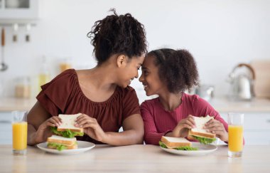 Mutlu Afro-Amerikan anne ve kızı evde birlikte vakit geçiriyorlar, öğle yemeği yiyorlar, taze portakal suyu içiyorlar, sandviç yiyorlar, alınlarıyla birbirlerine dokunuyorlar ve gülümsüyorlar.