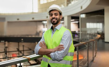 Happy confident young attractive arabic engineer in hard hat and protective clothing in office plant interior, free space. Professional work in industry, builder checking job at modern factory indoor