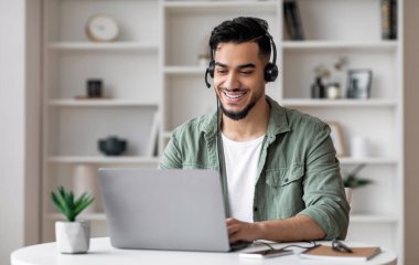 Kulaklıklı, sakallı, yakışıklı, yakışıklı, genç bir adam oturma odasındaki dizüstü bilgisayara bakıyor. Uzaktan danışmanlık, sosyal mesafe, covid-19 karantinası sırasında yeni normal ve müşteri desteği