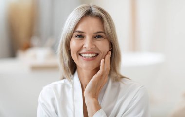 Closeup photo of attractive blonde middle aged woman in white silky bathrobe touching her pretty face and smiling, enjoying her morning face care routine, bathroom interior, copy space