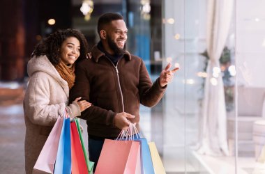 Satış ve Tüketim Konsepti. Gülümseyen Afro-Amerikan çift alışveriş merkezinin penceresine bakıyor ve işaret ediyor, akşamları alışveriş merkezinin yakınında yürüyorlar. Mevsimlik Satış ve İndirim, Satın Alım ve Perakende