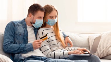Sadece online alışveriş. Koruyucu maskeli genç çift evde dizüstü bilgisayar ve kredi kartıyla yemek sipariş ediyor.