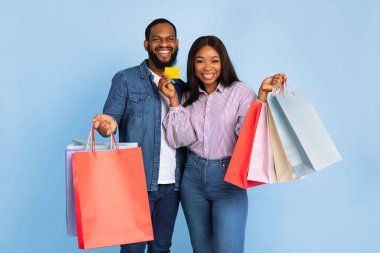Kolay ödeme. Gülümseyen siyah bir çiftin portresi alışveriş merkezinde alışveriş yaptıktan sonra alışveriş torbalarını tutan kredi kartını gösteriyor, turkuaz mavi stüdyo arka planında izole bir şekilde poz veriyor.
