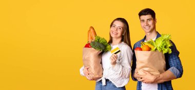 Modern alışveriş ve modern teknolojiyle temassız ödeme. Mutlu genç adam ve kadın ellerinde alışveriş poşetleri ve kredi kartıyla, sarı arka planda izole edilmiş, stüdyo çekimi, boş alan