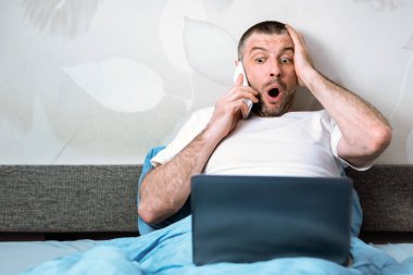 Şok olmuş, olgun bir adam cep telefonuyla konuşuyor. Coronavirus karantinası sırasında laptopta şok edici haberler okuyor..