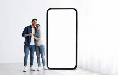 Genç Müslüman bir çift yan yana durur, neşeyle yanyana ışıldarken kapalı bir alanda içi doğal ışık dolu büyük, boş bir akıllı telefon ekranına yaslanır..