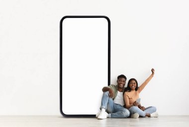 Neşeli siyah bir çift yerde oturuyor, rahatlamış ve gülümsüyor. Büyük, boş bir telefon ekranına yaslanırlar, birlikte geçirdikleri zamanın tadını çıkarırlar ve sohbet ederler..