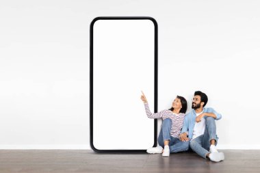 Genç Hintli bir çift yerde oturuyor, gülümsüyor ve boş bir akıllı telefon ekranını işaret ediyor. Minimum dekorlu modern bir odadalar, rahat bir atmosfer yaratıyorlar..