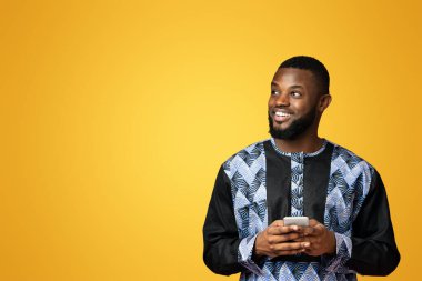 Modern akıllı telefonlu mutlu siyahi adam boş alana bakıyor, Afrika tişörtü giyiyor, pembe stüdyo geçmişi var. Geleneksel kostümlü Afrikalı Amerikalı genç adam cep telefonu ve panorama kullanıyor.