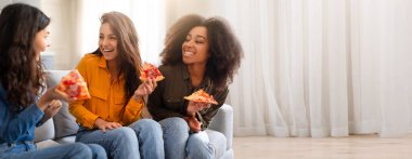 Üç çok ırklı arkadaş gülüyor ve sıcak bir oturma odasında pizza dilimlerini paylaşıyorlar. Onların neşeli ifadeleri dostluk ve eğlenceyle dolu bir toplantıyı vurguluyor. Mekan parlak ve davetkar..