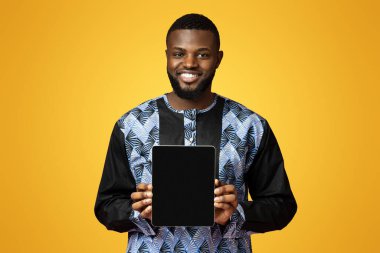 Siyah neşeli adam elinde boş ekranlı dijital tablet tutuyordu. Geleneksel Afrika kostümü giyiyordu. Pozitif Afro-Amerikalı genç adam pembe stüdyo arka planında yeni bir uygulama gösteriyor.