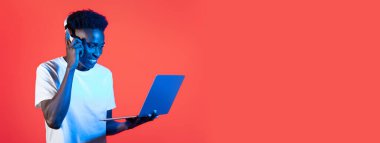 Beyaz tişörtlü Afro-Amerikalı genç bir adam dizüstü bilgisayar kullanırken telefonla konuşuyor. Gülümsüyor, işine dalmış kırmızı bir zemine karşı..