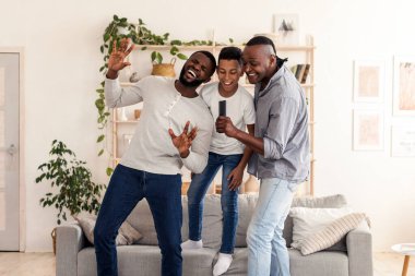 Aile eğlencesi. Neşeli siyah baba, oğul ve büyükbaba evde birlikte takılıyor, birlikte şarkı söylüyor ve eğleniyorlar, uzaktan kumandayı mikrofon olarak kullanıyorlar.