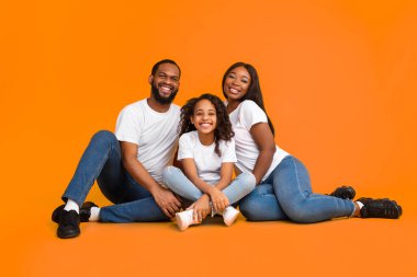 Aile Günü, Ebeveynlik ve Çocukluk Konsepti. Gülümseyen genç siyahi anne-baba, çocuk yaşta yerde oturan baba, sarılan kız, sarı turuncu arka plan stüdyo resminde izole edilmiş.