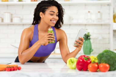 Siyah bir kadın telefonda mesajlaşıyor ve ev yapımı detoks suyu içiyor spor giysiler giyiyor, mutfak masasına yaslanıyor.