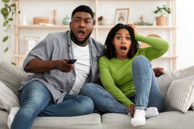 Coronavirus Haberleri. Şok olmuş Afro-Amerikan çift, açık ağızla televizyon izliyor, birlikte evde oturuyor, oturma odasında kanepede oturuyor,