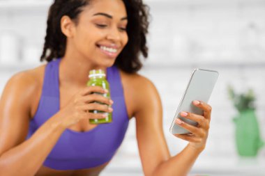 Siyah kızın cep telefonuna baktığı, şişeden yeşil sağlıklı sebze suyu içtiği, cihaza odaklandığı yakın plan.