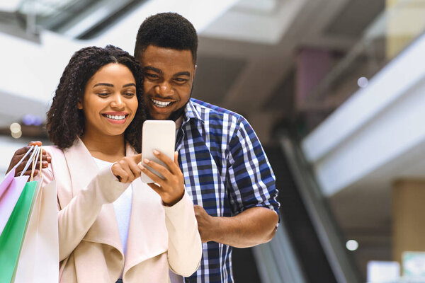 Скидка App. Happy Black Couple Using Smartphone In Shopping Mall, Checking Sale Price Offers, Received Cashback Bonus for Purchases, Free Space
