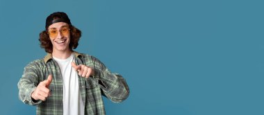 Güneş gözlüklü genç adamın portresi turkuaz arka planda seni işaret ediyor.