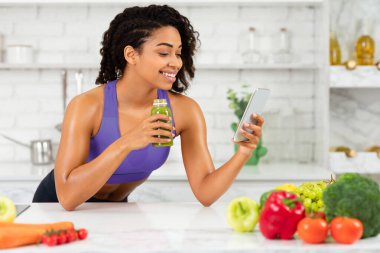 Günaydın Smoothie. Masaya yaslanan, telefon kullanan ve mutfakta ev yapımı detoks suyu içen Afrikalı kadın portresi.