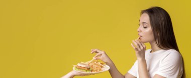 Fast Food Bağımlısı. Genç bayan yemek yiyor sağlıksız yemekle sunulan tabaktan patates kızartması alıyor, sarı arka planda duruyor, stüdyoda boş yer var.