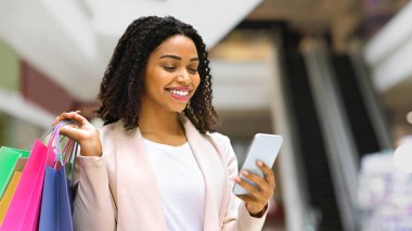 İndirimli uygulama. Gülümseyen Afrikalı Amerikalı Kadın alışveriş merkezinde akıllı telefon kullanıyor, indirimleri kontrol ediyor, bedava yer açıyor.