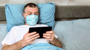 Coronavirus Karantinası. Maskeli orta yaşlı adam dijital tablet kullanıyor. Evde yatakta uzanmış film seyrediyor. Seçici Odaklanma
