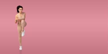 Genç bir kadın, parlak pembe bir stüdyo ortamında spor yaparken diz kaldırma egzersizi yapıyor. Odaklanmış ve rahat bir idman kıyafeti giyiyor..