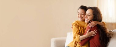 Parlak, havadar bir oturma odasında bir anne ve kızı sevgi dolu bir kucaklaşmayı paylaşırken neşeyle gülümserler. Evlerinin rahatlığı bu sevgi dolu anı güçlendiriyor..