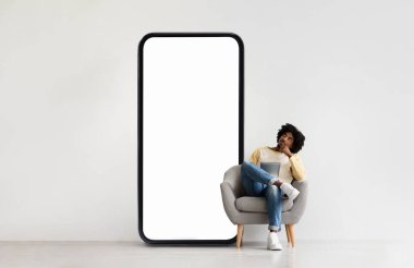 Bir insan, modern bir sandalyede, dev bir akıllı telefon şeklindeki ekranın yanında düşünürken rahatlar. Kapalı mekanın minimalist bir tasarımı ve nötr tonları var..
