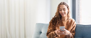 Akıllı telefonuna mesaj atarken kıvırcık saçlı genç bir kadın gülümsüyor, parlak bir odada rahat bir kanepede oturuyor. Arkadaşlarıyla yakınlaşırken doğal ışık onun neşeli yüz ifadesini arttırır..